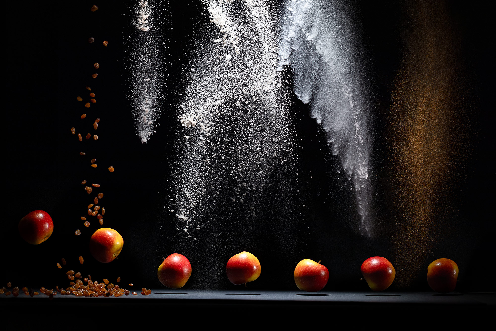Ingredients for apple pancakes recipe. The image shows an apple running across a black table with black backdrop. Raisins, salt, flour, baking soda and cinnamon powder are falling from above.