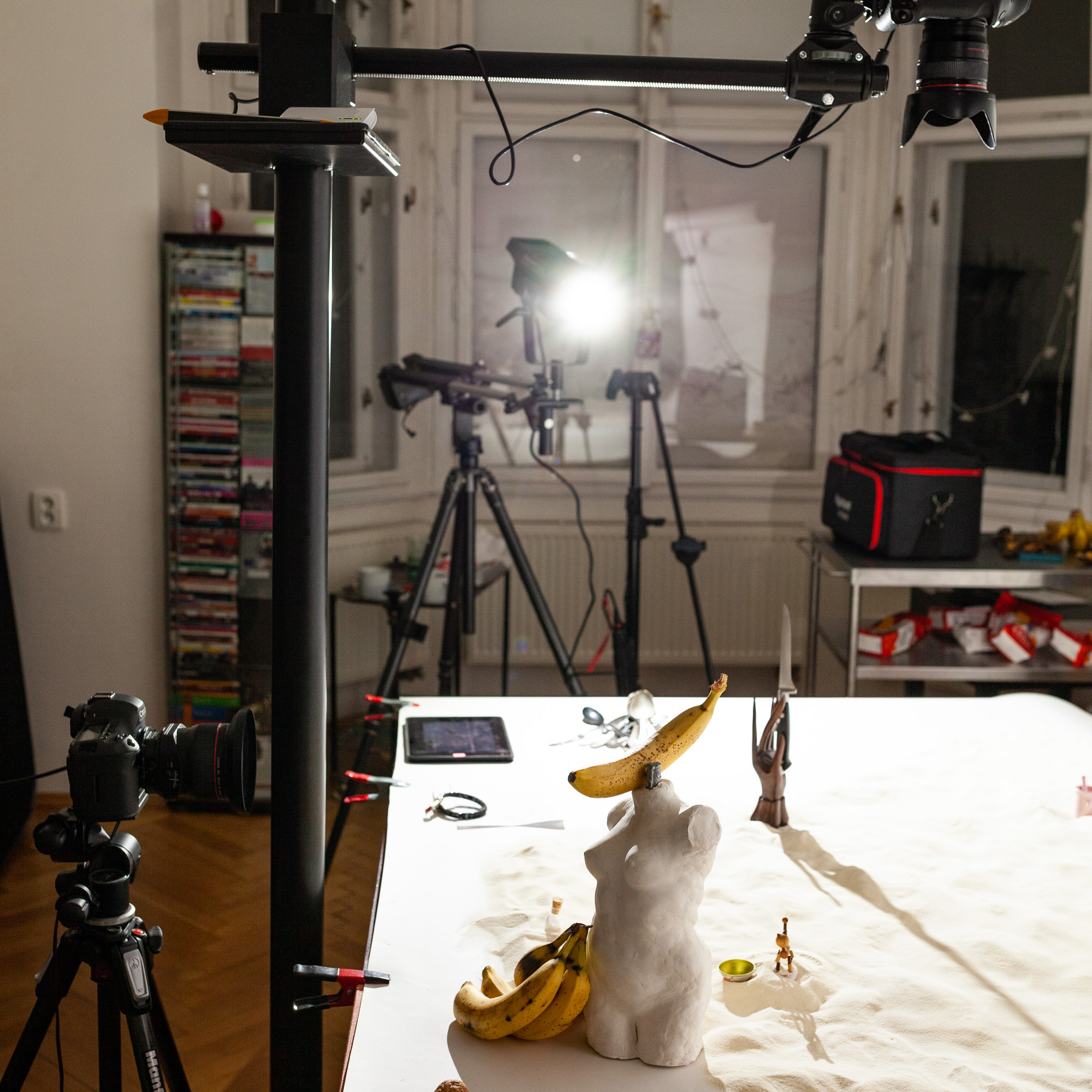 Behind the scenes of foodlydoodlydoo's vegan banana bread recipe. The photo shows the preparation of a scene. A statue of a naked women's torso decorated with bananas is seen as well as a wooden hand holding a knife. Cameras and lights are set around the scene.