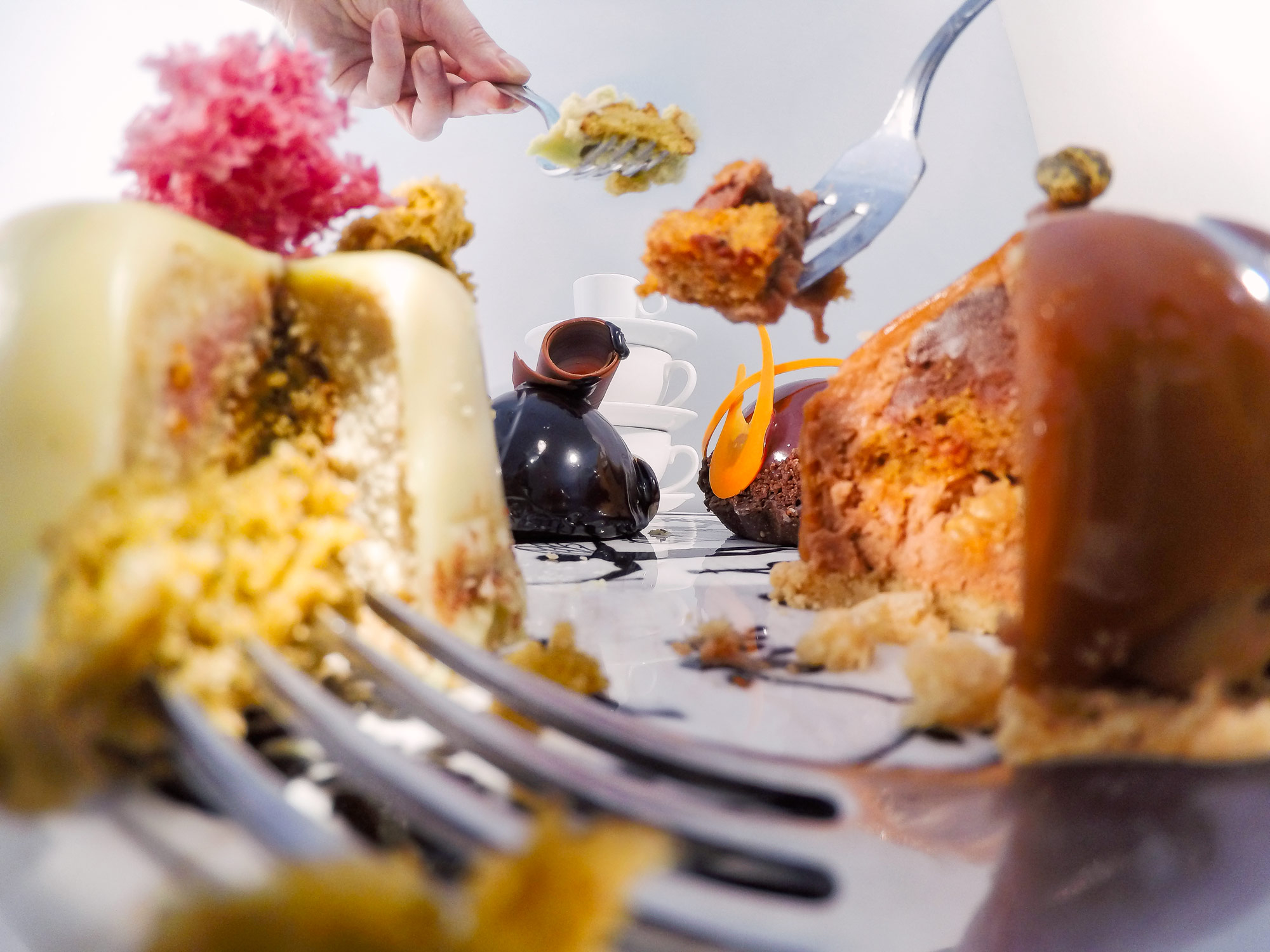 Inside food photo series, depicting a close up view of cakes and forks. From the point of view of the cake as standing on a table in the middle of a cake party, surrounded by forks, cakes and cups with chunks of cakes being forked through the air.