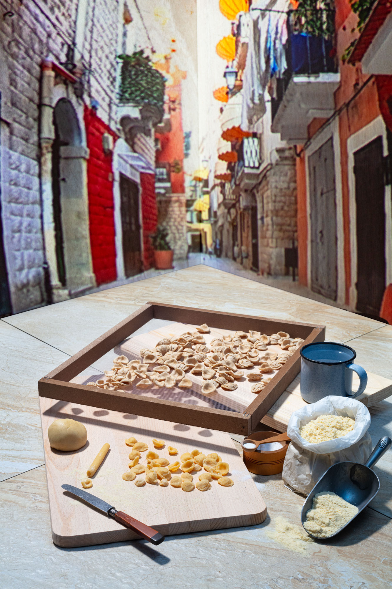 A view of a typical Italian narrow old town street in Apulia. Traditional Orecchiette pasta is layed out to dry on a grid. A bag of flour, salt and a cutting board lying next to it. 