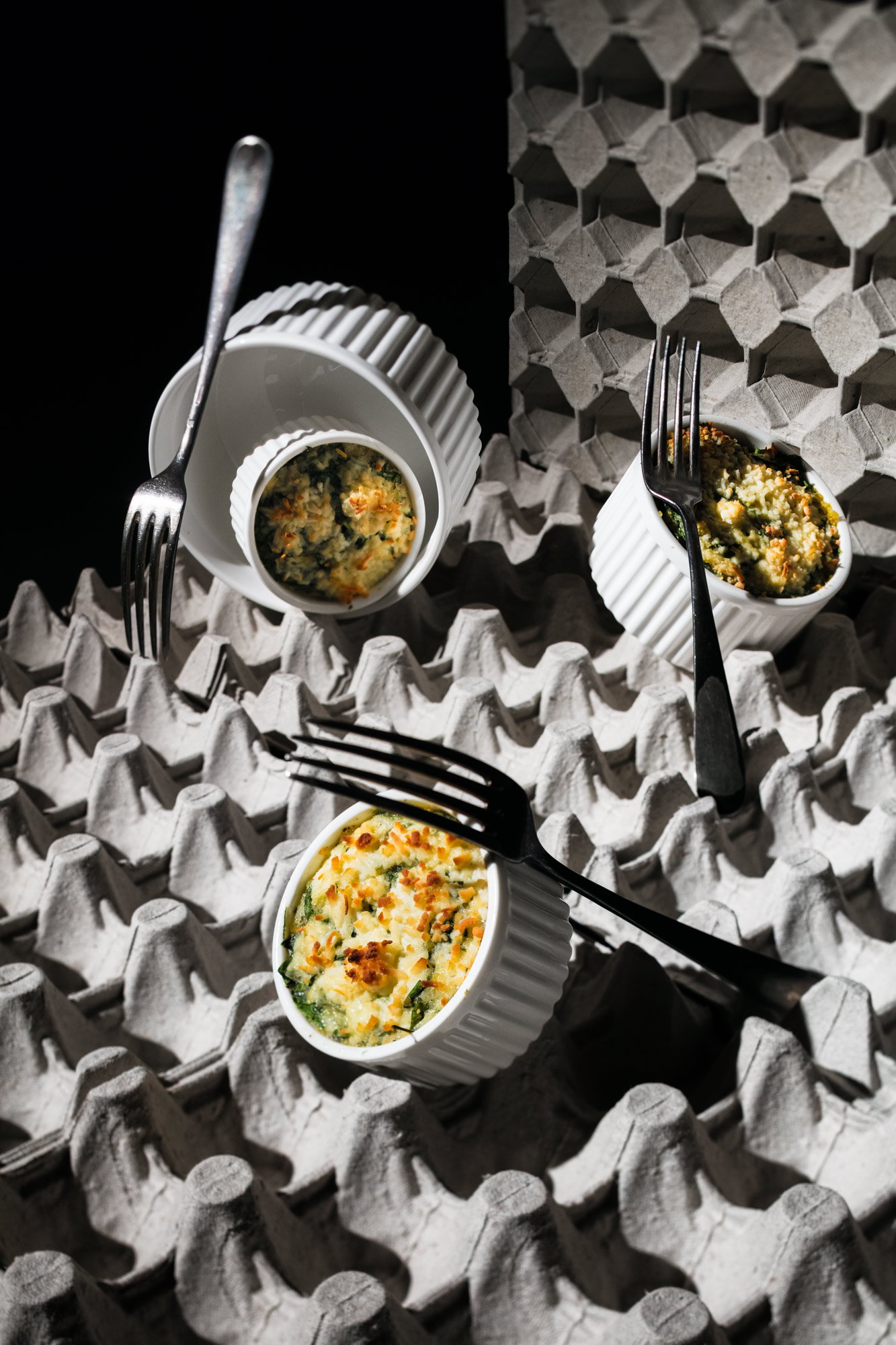 Four round casserole dishes and three forks displayed on two adjusting egg cartons. three of the dishes are filled with a spinach millet casserole. Depicted in a harsh contrast light with deep shadows